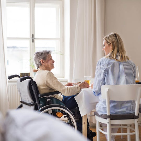 A senior woman in wheelchair with a health visitor sitting at the table at home.